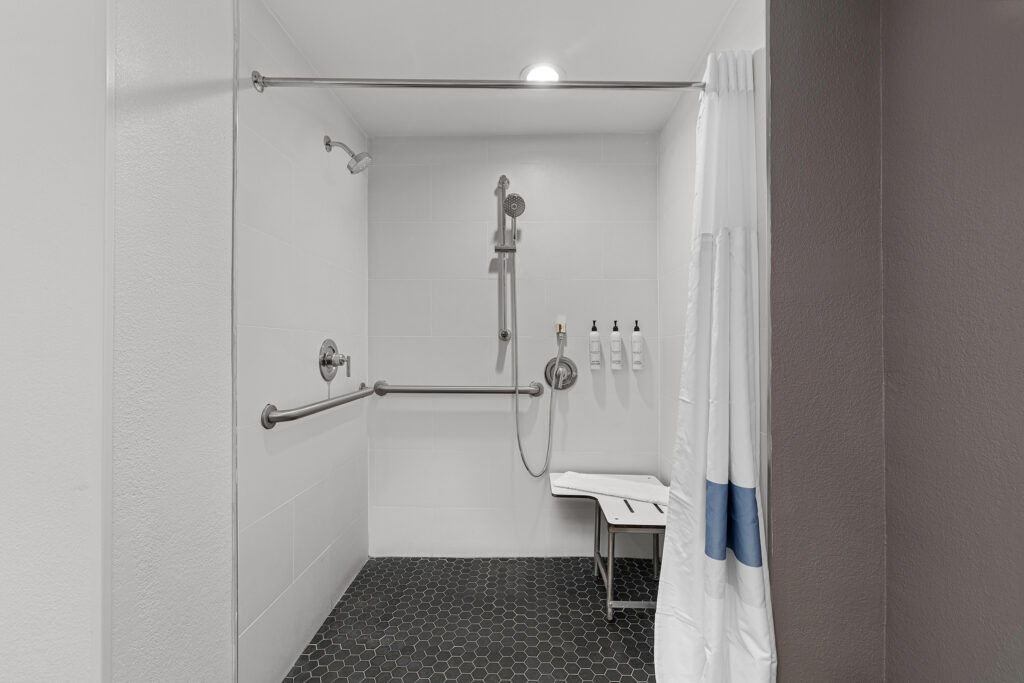 Accessible walk-in shower at the Marriott Yulee hotel features a wall-mounted showerhead, grab bars, foldable seat, dark hexagonal floor tiles, white wall tiles, three wall-mounted soap dispensers, and a partially visible shower curtain.