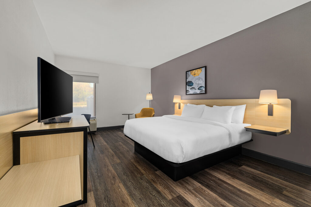A modern hotel room in Yulee, Florida, featuring a large bed with white bedding, wooden floors, a wall-mounted headboard with lamps, a TV on a wooden table, an armchair, desk, and a large window letting in natural light.