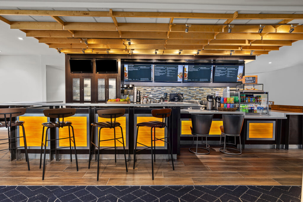 A modern cafe bar with high chairs, illuminated yellow panels, digital menu boards, and a wood-panel ceiling. Perfect for hospitality photography, the setting is clean, bright, and welcoming—ideal for any hotel or courtyard space.