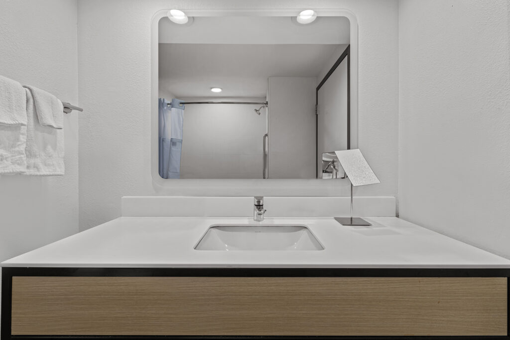 Modern bathroom in a Yulee hotel with a rectangular sink, minimalist faucet, mounted mirror, towel rack with white towels on the left, and a small angled mirror on the right side of the counter. Shower area visible in the background—perfect for your Florida stay.