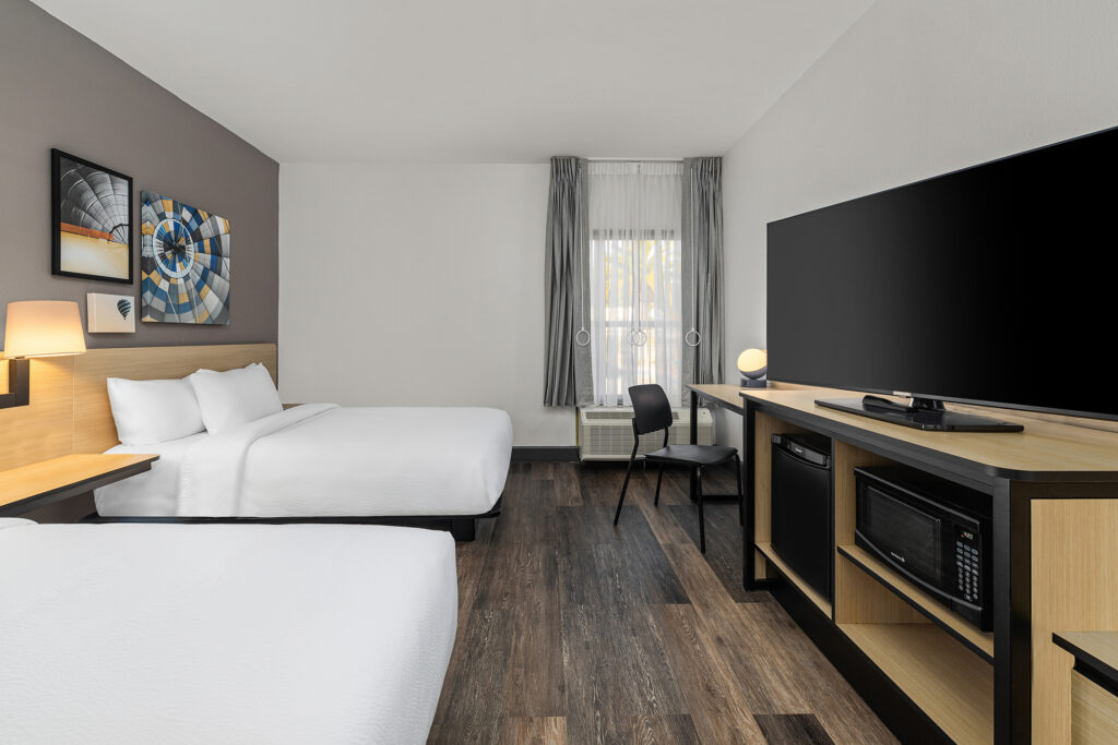 Modern Marriott hotel room in Florida featuring two double beds, a large flat-screen TV on a wooden unit with microwave and mini-fridge, a desk by the window, and abstract artwork above the beds.