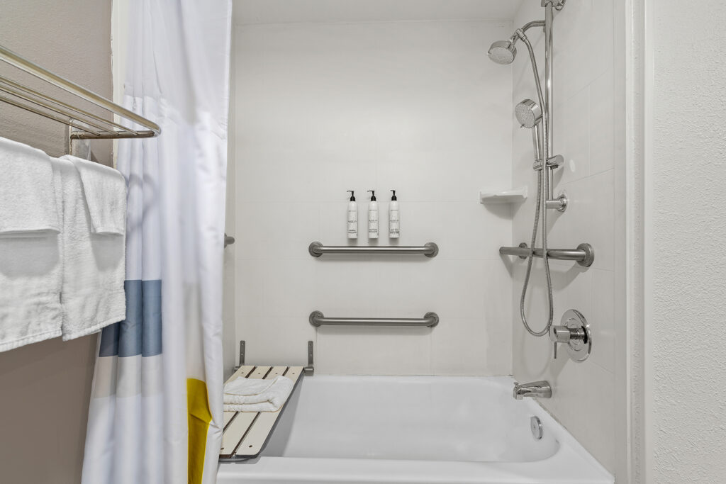 Accessible bathtub with grab bars, showerhead, bath bench, and three toiletry dispensers mounted on a white wall at Marriott in Yulee, Florida. Towels rest on a rack and a shower curtain is partially open on the left.