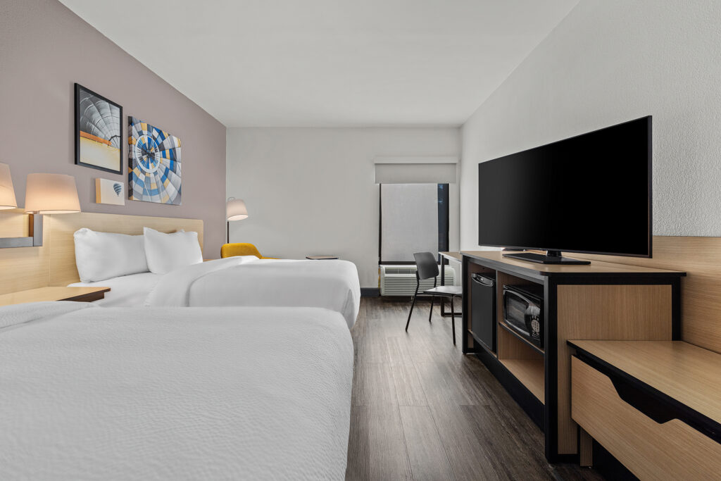 A modern hotel room at City Express with two double beds, a flat-screen TV on a wooden stand, a work desk with chair, wall art above the beds, and a large window with blackout shade. The room features wood flooring and neutral decor.