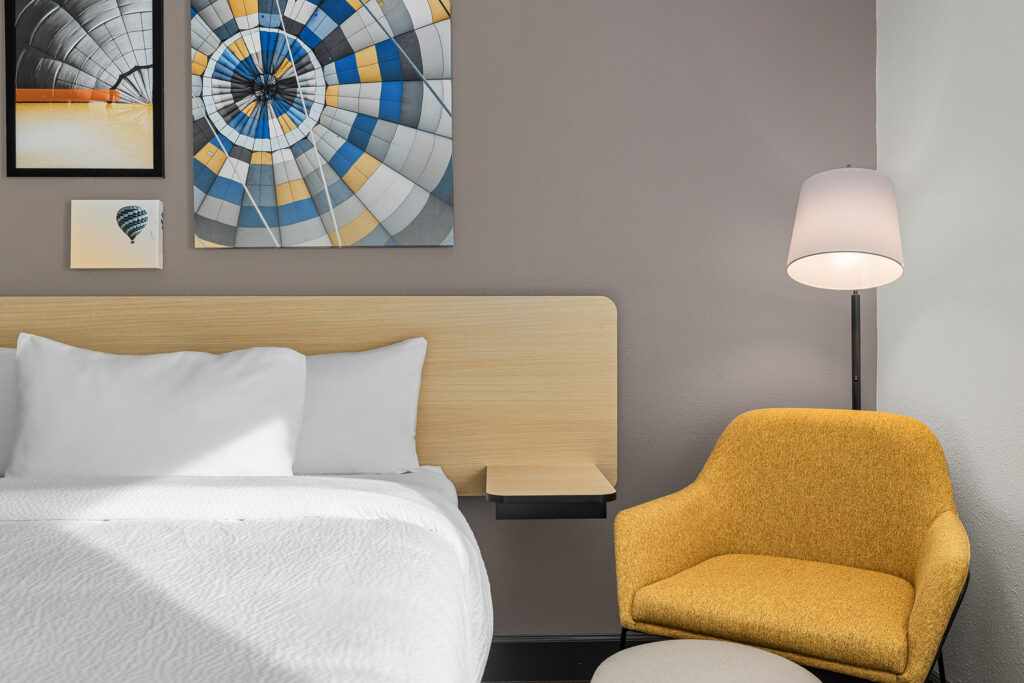 A modern Marriott hotel room in Florida featuring a white bed, yellow armchair, round ottoman, floor lamp, and wall art of hot air balloons above the bed on a gray accent wall.