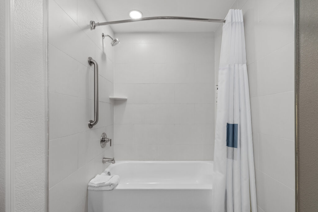 A white tiled bathtub and shower combo with a curved shower rod, white curtain, small corner shelf, grab bar, and a neatly folded white towel on the tub edge—perfect for a relaxing stay at a City Express hotel in Florida.