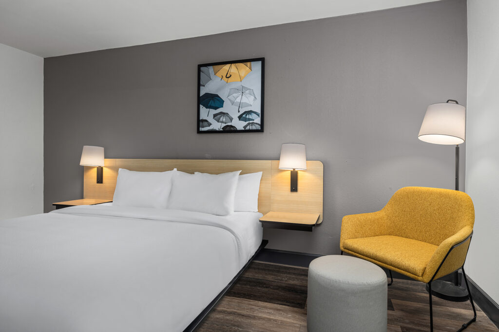 A modern City Express hotel room in Yulee, Florida, featuring a neatly made bed, wall-mounted lamps, a yellow armchair, a round gray ottoman, a floor lamp, and a framed picture of umbrellas on a gray accent wall.