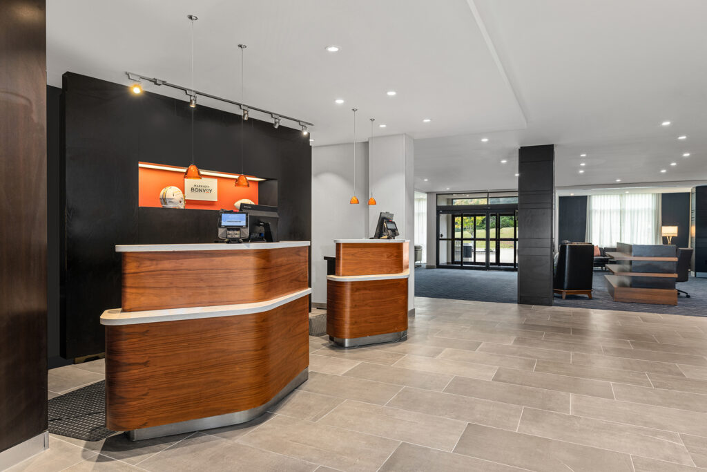 A modern hotel lobby in Oxford, MS with a wooden reception desk, pendant lights, tiled floor, and a courtyard seating area in the background. The space is clean, well-lit, and showcases stylish hotel photography and contemporary decor.