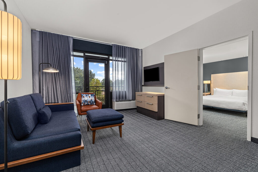 A modern hotel suite in Oxford, MS, featuring a blue sofa, brown leather chair, and ottoman. Large windows reveal a balcony with courtyard views, perfect for hospitality photography. A flat-screen TV is mounted above the dresser; the bedroom offers a king bed.