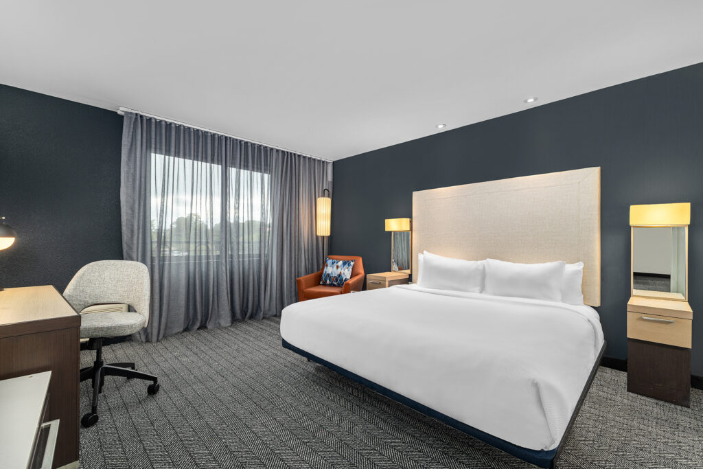 Modern hotel room in Oxford, MS, with a large bed, white bedding, two nightstands and lamps, an orange armchair, desk and chair, plus a large window with sheer curtains—perfect for hotel photography or hospitality photography.