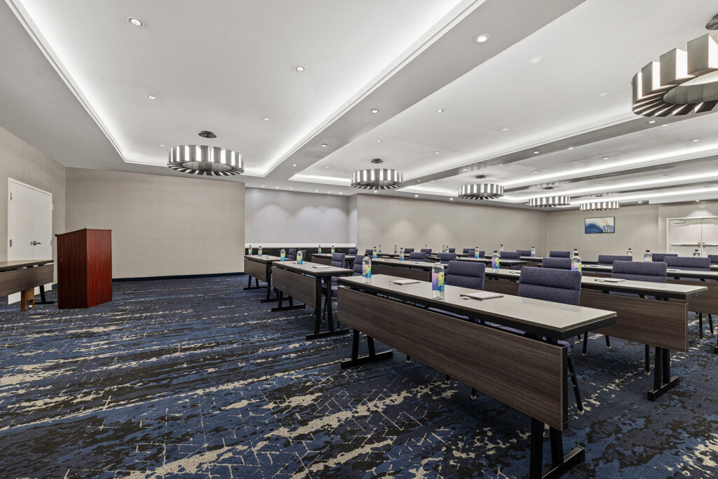 A modern conference room at Marriott with rows of tables and chairs, each set with water bottles, glasses, and notepads. Perfect for hotel photography, the space features a blue patterned carpet, neutral walls, and contemporary ceiling lights.