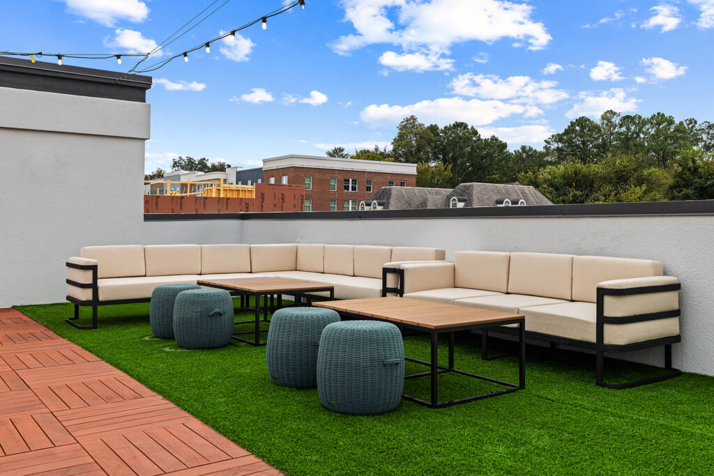Modern rooftop patio at Marriott with cream-colored sofas, wooden tables, and blue woven ottomans on green artificial grass. String lights hang above, and buildings and trees are visible in the background under a blue sky.