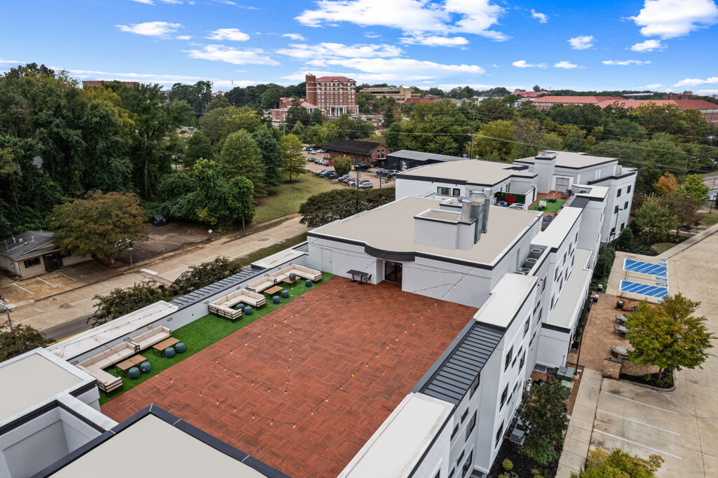 Aerial view of a modern building in Oxford, MS with a spacious rooftop terrace featuring seating areas, greenery, and tables. The hospitality photography captures surrounding trees, a courtyard, and other buildings under a partly cloudy sky.