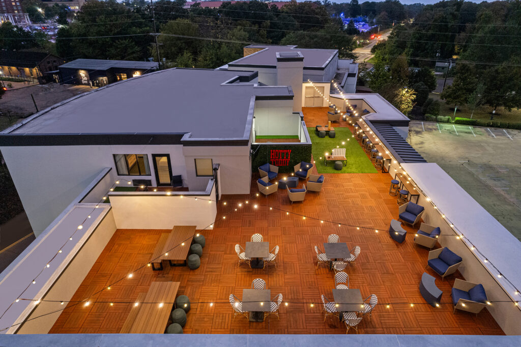 A rooftop patio at dusk with string lights, various seating areas, lounge chairs, greenery, and wooden flooring—perfect for hotel photography. This Marriott Courtyard outdoor space overlooks trees, buildings, and a parking lot.