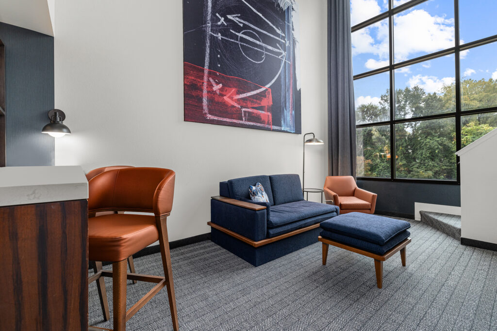 Modern living room with tall windows, a blue sofa, matching ottoman, orange chair, barstool, floor lamp, and abstract wall art. Sunlight streams in, highlighting the cozy décor and courtyard views—perfect for hospitality photography.