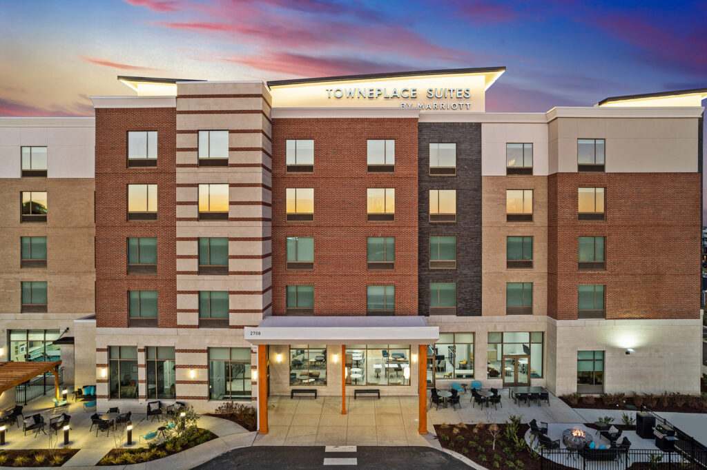 A modern four-story TownePlace Suites hotel in Murfreesboro at sunset, featuring a well-lit entrance, large windows, and inviting outdoor patio seating beneath a sky blending blue and pink hues.