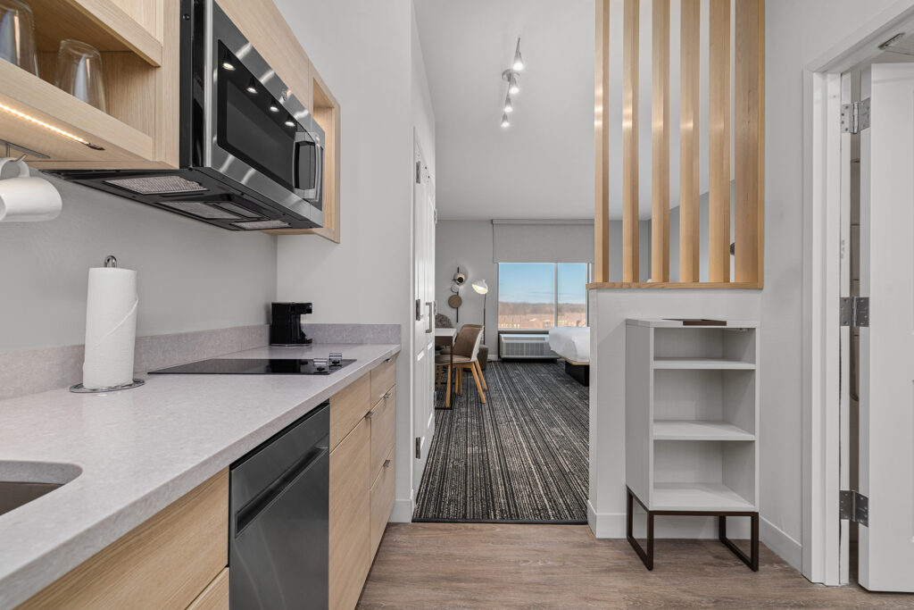 Modern studio apartment with a compact kitchen on the left, featuring a sink, stove, microwave, and dishwasher. Experience TownePlace Suites-style hospitality in TN, with light wood and white decor and a bedroom area with a large window.