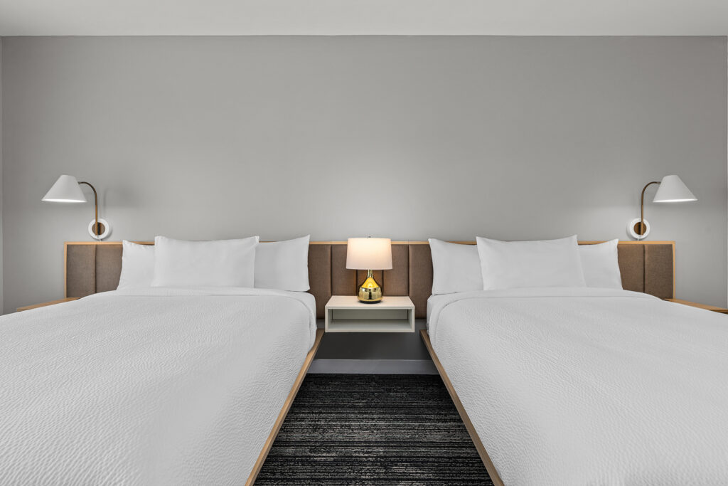 A TownePlace Suites hotel room in Murfreesboro, TN features two neatly made double beds, a modern nightstand with a lamp between them, and wall-mounted reading lights above each bed against a light gray wall.