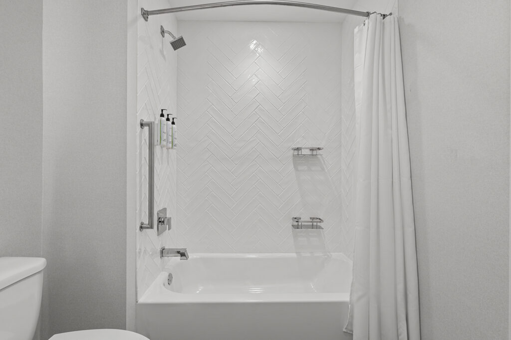 A modern white bathroom at TownePlace Suites features a bathtub, showerhead, wall-mounted toiletries, two metal shelves, and a white shower curtain. The wall above the tub showcases a striking herringbone tile pattern.