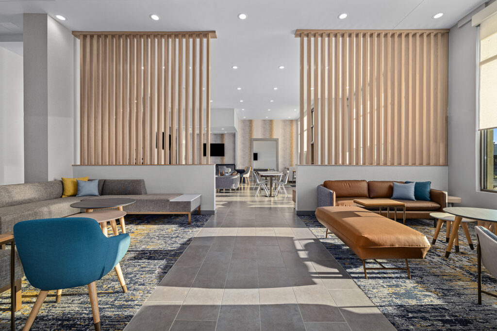 A modern lounge area at TownePlace Suites by Marriott features neutral tones, gray and brown sofas, blue chairs, wooden slat dividers, patterned carpet, and abundant natural light from large windows. Tables and chairs are arranged in the background.