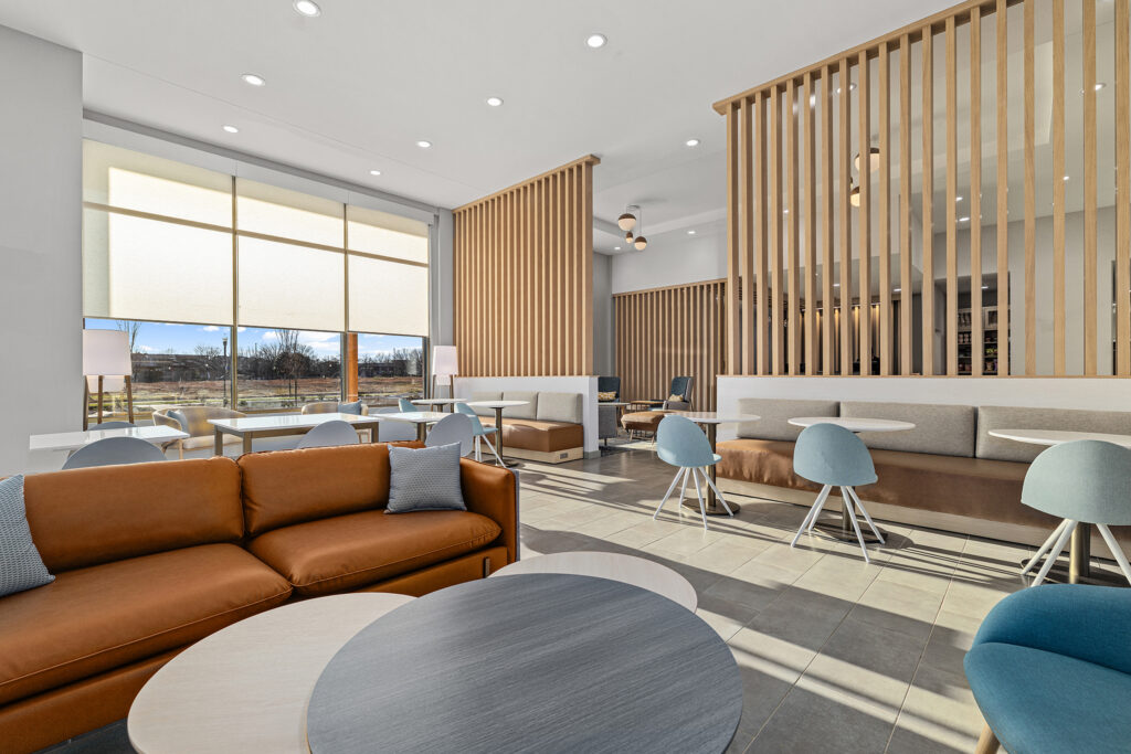 A modern, sunlit lounge at TownePlace Suites Murfreesboro features brown sofas, light blue chairs, round tables, and wooden slat dividers. Large windows fill the hotel space with natural light for a bright, welcoming atmosphere.