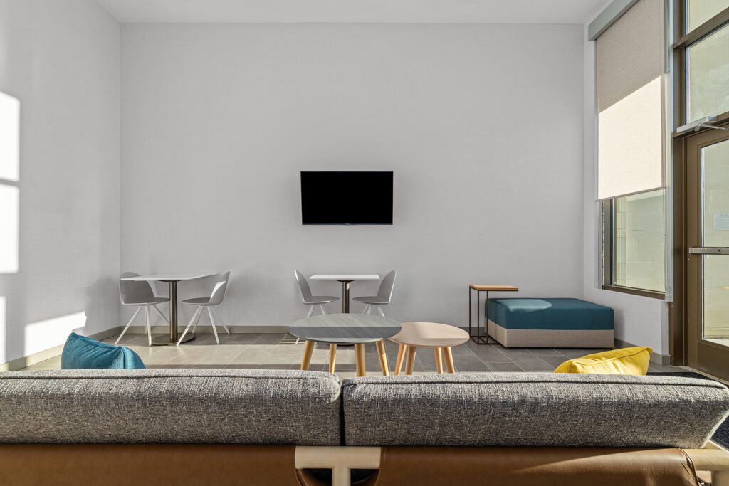 A modern lounge room at TownePlace Suites hotel in TN features a gray sofa, colorful cushions, two small round coffee tables, sets of white chairs and tables, a wall-mounted TV, and large windows flooding the space with natural light.