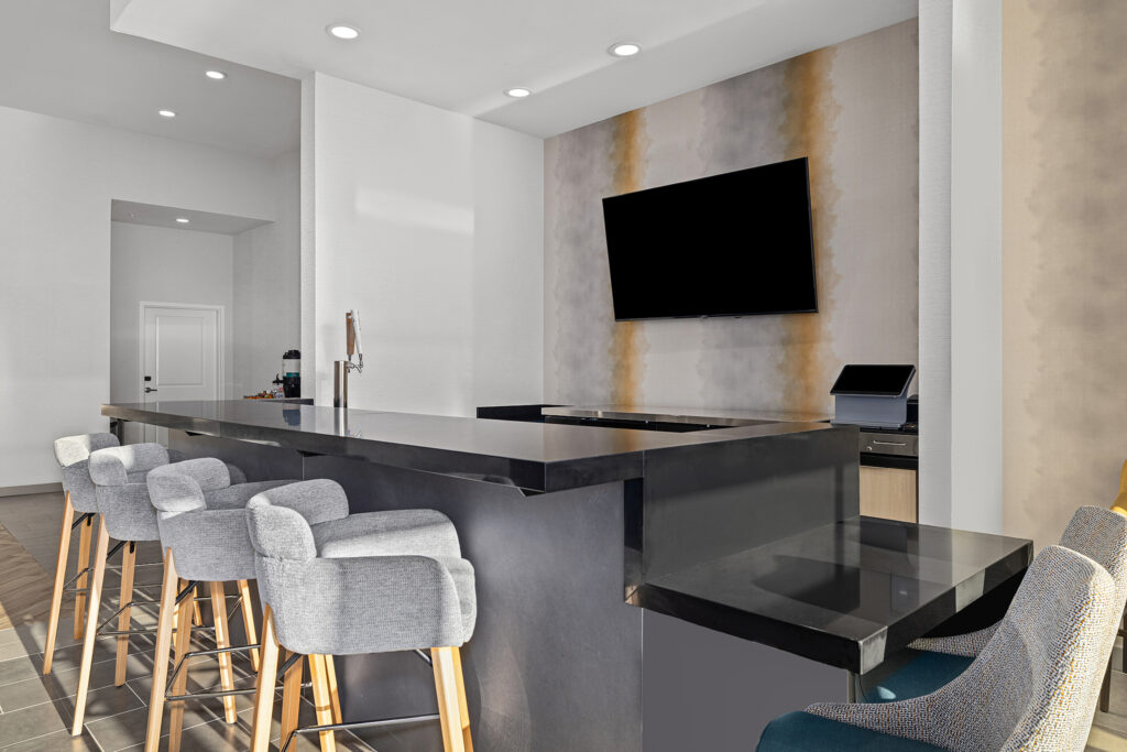 Modern bar area at TownePlace Suites Murfreesboro, featuring a sleek black countertop, five gray upholstered barstools with wooden legs, a wall-mounted flat-screen TV, and minimalist Marriott-inspired decor in neutral tones.