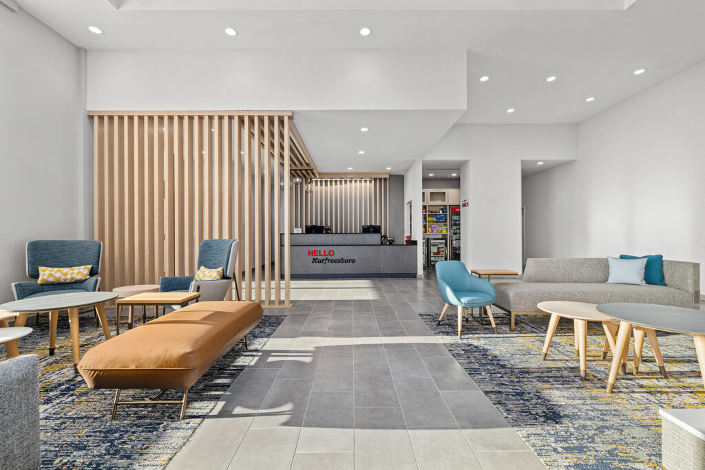 Modern Marriott hotel lobby in TN with contemporary furniture, wooden slat room divider, reception desk at the back, gray tiled floor, and patterned carpet accents creating a bright, welcoming atmosphere.