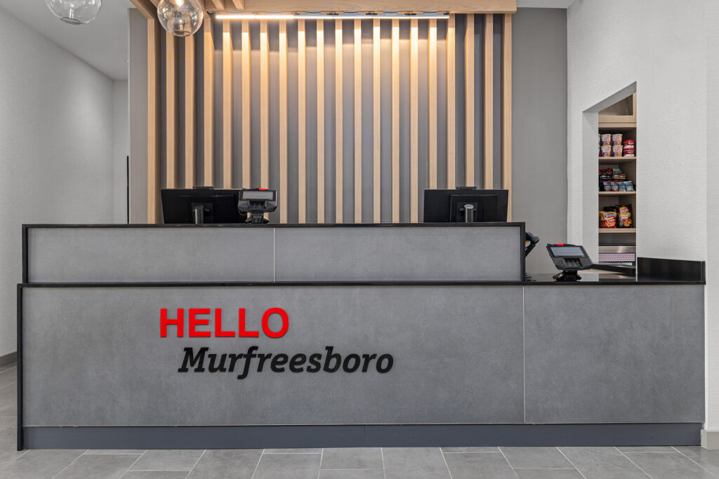 A modern TownePlace Suites hotel reception desk with HELLO Murfreesboro on the front, two computers, pendant lights above, and a small snack shelf to the right. Vertical wooden slats decorate the back wall.