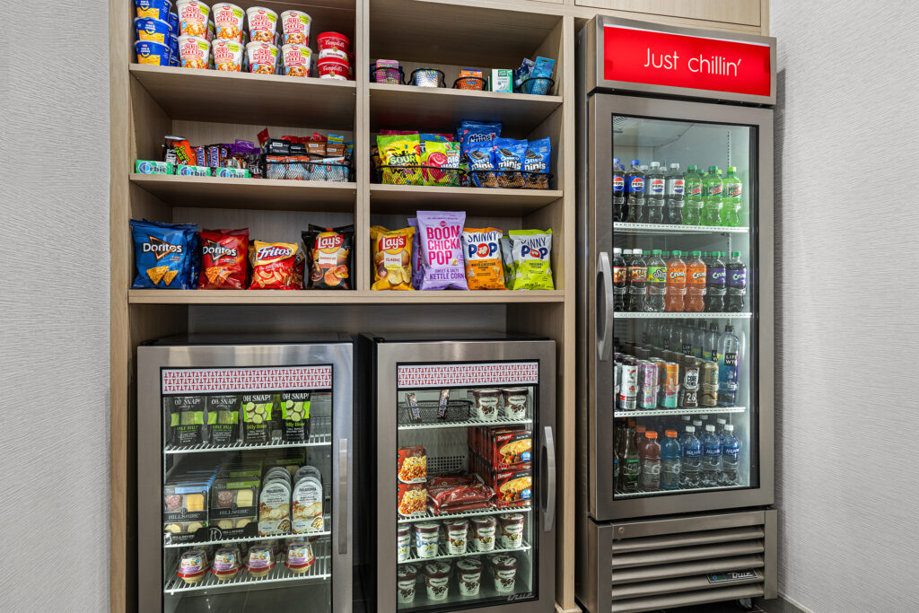 Snacks and drinks in the TownePlace Suites Marriott Murfreesboro market area, with shelves of chips, instant noodles, and candy, plus three refrigerators stocked with bottled drinks, sandwiches, yogurt, fruit, and sodas. A red Just chillin sign hangs above.