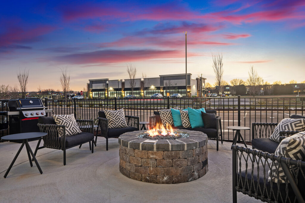 Outdoor patio at sunset at the Marriott Murfreesboro, with cushioned chairs around a circular stone fire pit. Decorative pillows add color, and a barbecue grill sits nearby. Modern hotel buildings and trees stand against the vibrant sky.