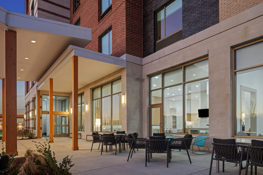 Modern building exterior at TownePlace Suites Murfreesboro, TN, featuring large windows, a covered patio area, and outdoor seating with black woven chairs and tables. The well-lit patio offers views into the inviting interior through the windows.