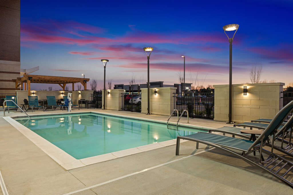 Outdoor pool area at sunset at TownePlace Suites by Marriott TN, with lounge chairs, pool ladders, and lit street lamps; a pergola and fence enclose the modern space under a vibrant pink and blue evening sky.