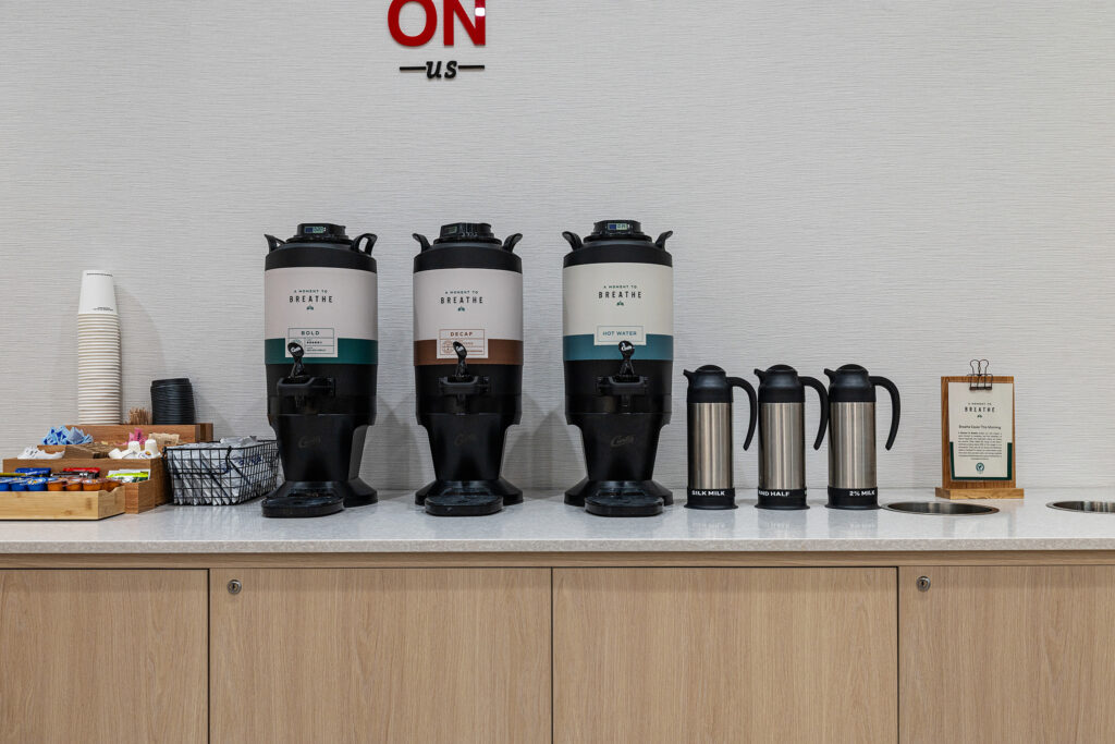 Three large coffee dispensers labeled “DECAF” and “BREWED” sit on a counter in Murfreesboro, TN, surrounded by coffee pots, cups, sweeteners, stirrers, and lids—a welcoming touch of hospitality against a plain background wall.