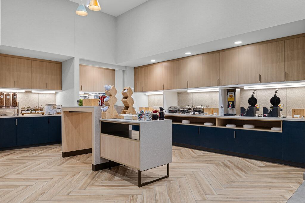 Modern hotel breakfast buffet area at TownePlace Suites Marriott Murfreesboro features wood flooring, blue and light wood cabinets, breakfast items, cereal dispensers, coffee machines, and pendant lights in a clean, well-lit setting.