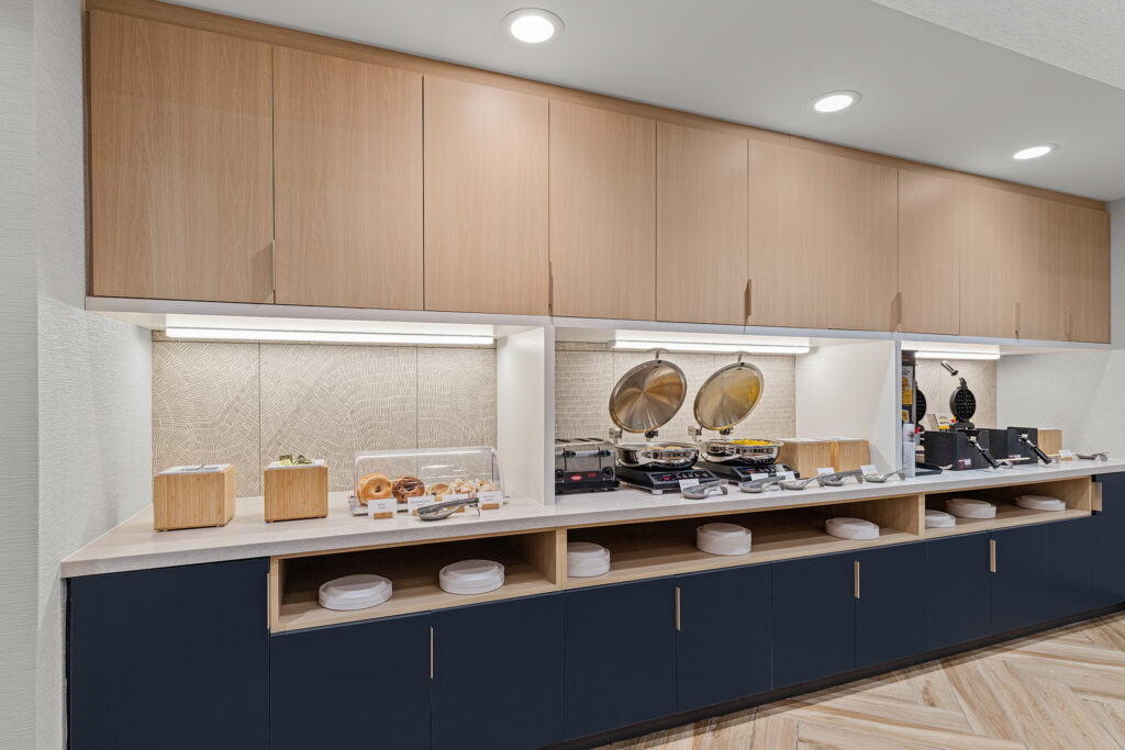 A hotel breakfast buffet with wooden cabinets, serving trays of pastries, chafing dishes of hot food, a waffle maker, and neatly stacked plates reflects Marriott’s signature hospitality and inviting atmosphere.