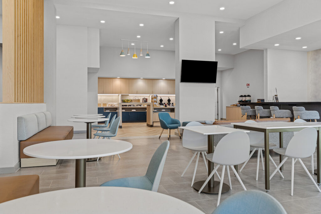 Modern Murfreesboro hotel breakfast area with white tables, blue and gray chairs, a booth, and a buffet counter in the background. Pendant lights hang from the ceiling, and a TV is mounted on the wall in this bright Marriott TN space.