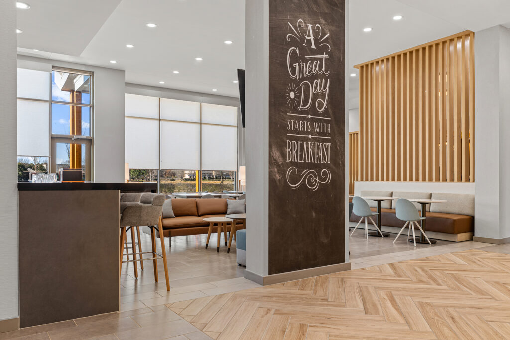 Modern hotel dining area at TownePlace Suites Marriott Murfreesboro with light wood floors, neutral furnishings, large windows, and a decorative pillar reading A Great Day Starts With Breakfast. Seating includes chairs, booths, and a communal table.