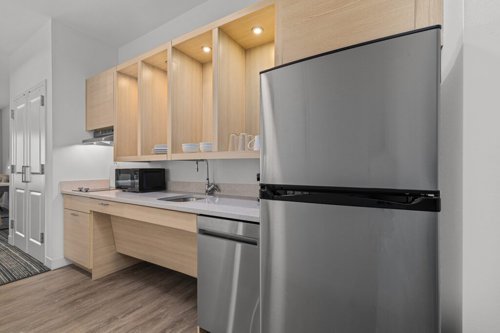 Modern kitchen at TownePlace Suites in TN with light wood cabinets, open shelves with dishes, stainless steel refrigerator, microwave, sink, dishwasher, and electric stovetop on wood flooring. Bright, neutral lighting offers hospitality.