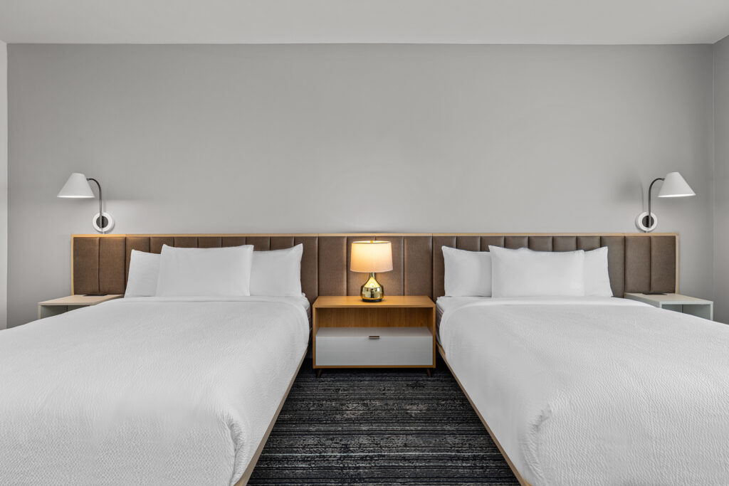Hotel room at Marriott Murfreesboro, TN, with two double beds, white bedding, a shared wooden headboard, a nightstand between them, a lit lamp, and two wall-mounted reading lights against a neutral gray wall.