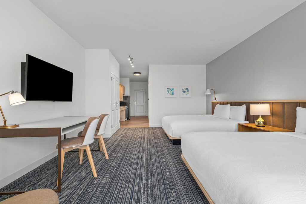 A modern TownePlace Suites hotel room in Murfreesboro, TN, features two double beds, a wall-mounted TV, desk with two chairs and a lamp, nightstands, framed art, and a kitchenette. The decor is minimalist and neutral.