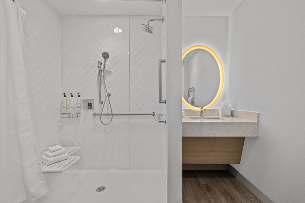 Accessible hotel bathroom at Marriott with a walk-in shower, grab bars, mounted showerhead, towels on a rack, and toiletry dispensers. Adjacent is a vanity with a sink and a round mirror with a glowing light, all reflecting true hospitality.