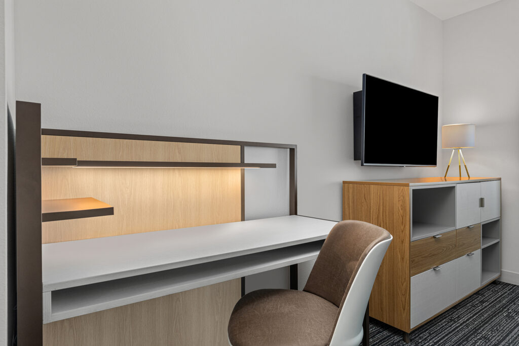 A modern workspace at TownePlace Suites Marriott Murfreesboro features a wooden desk and chair, under-shelf lighting, and a flat-screen TV above a cabinet with a lamp, all set against a light-colored wall.