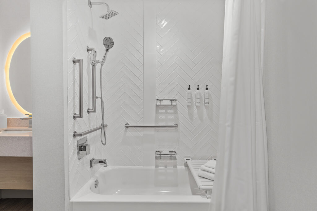Accessible bathroom at TownePlace Suites TN features a white bathtub with grab bars, handheld showerhead, and mounted soap dispensers. White shower curtain partially open; towels stacked on a rack inside the tub. Sink and mirror are visible on the left.
