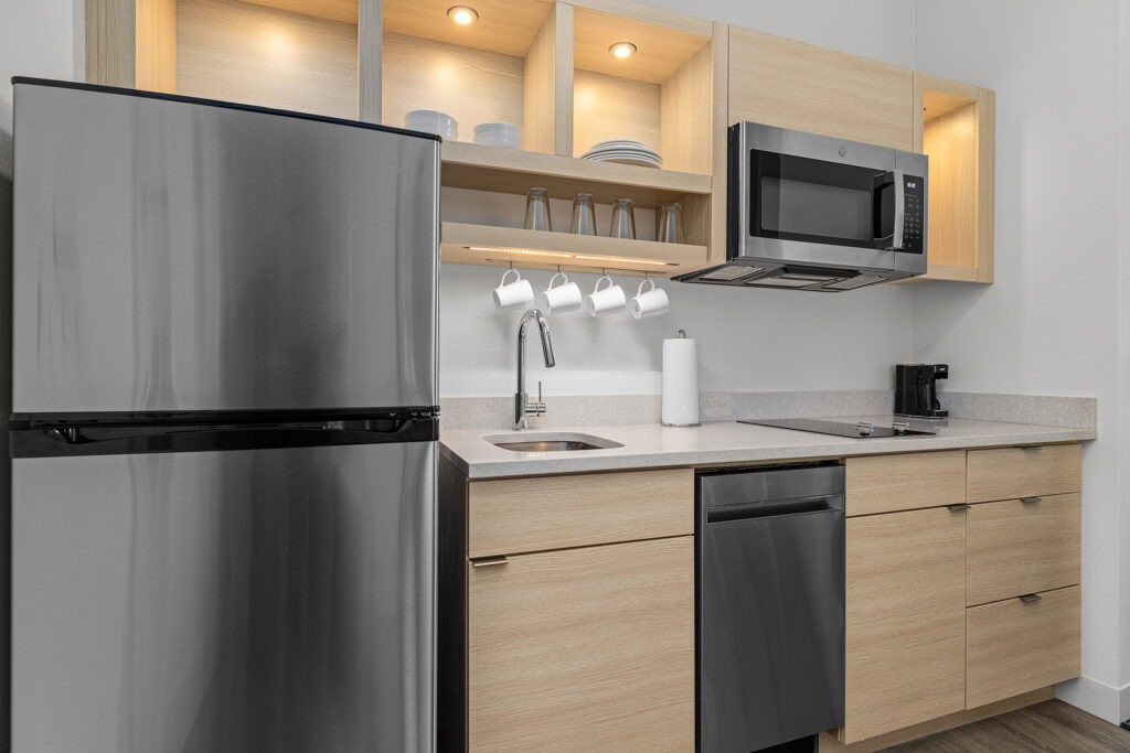 Modern kitchenette brings Marriott hotel hospitality home with a stainless steel refrigerator, dishwasher, microwave, and sink. Light wood cabinets hold dishes and glasses; enjoy coffee under warm under-cabinet lighting.