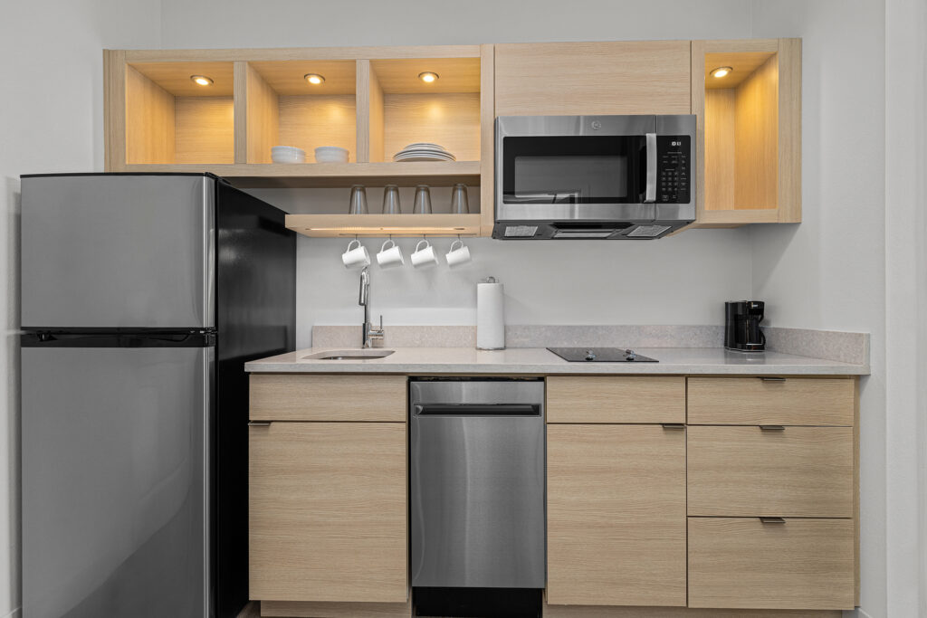 Modern kitchen at TownePlace Suites features light wood cabinets, stainless steel appliances including a refrigerator, microwave, and dishwasher, open shelves with dishes, an electric stove, and a small coffee maker on the countertop.