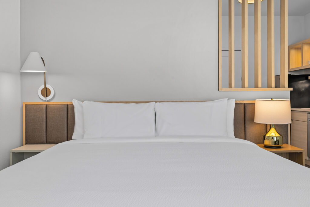 A neatly made bed with white linens and two pillows sits against a brown padded headboard at TownePlace Suites Marriott TN. A wall lamp is on the left and a table lamp on a nightstand to the right, with wooden panel accents in the background.