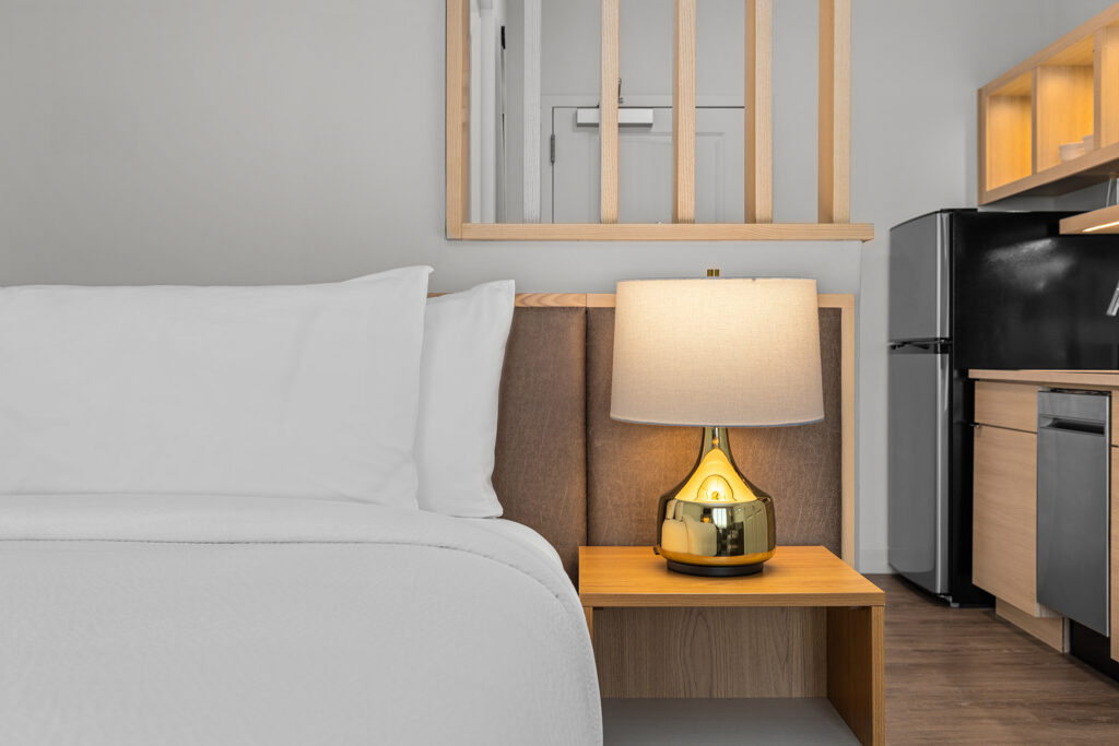 A neatly made bed with white linens beside a wooden nightstand holding a gold lamp with a white shade; a kitchenette and refrigerator are visible in the background at TownePlace Suites by Marriott in TN.