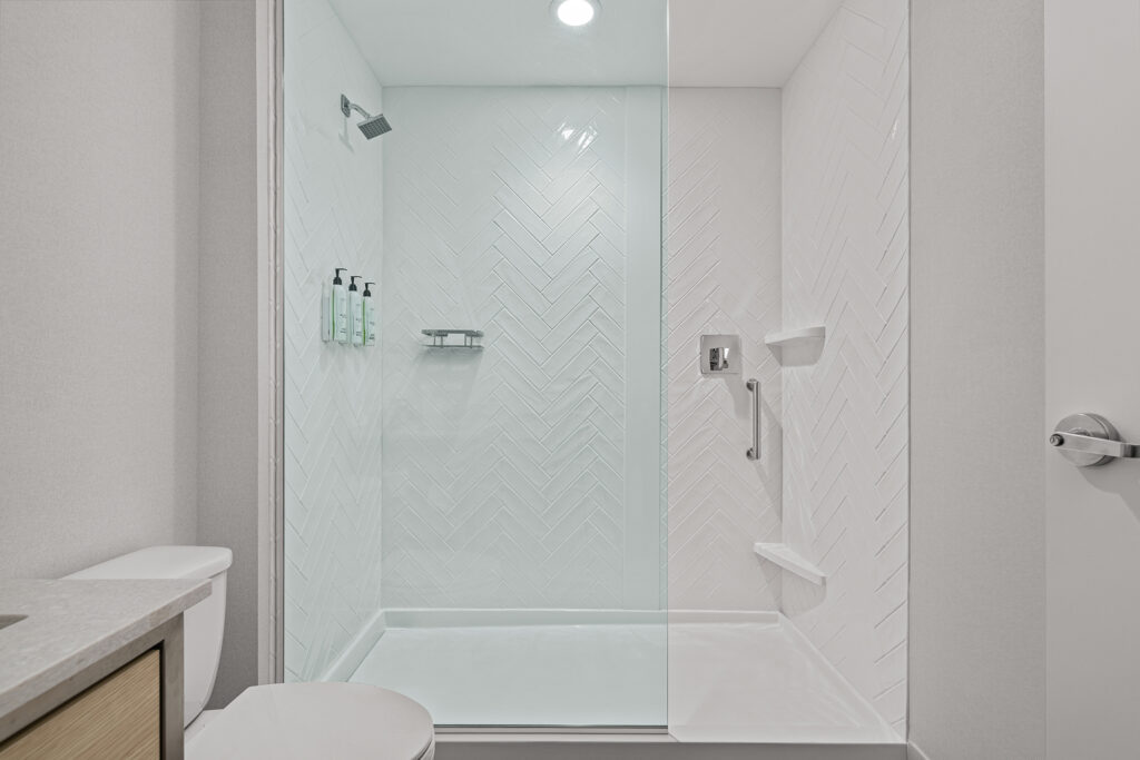 Modern, white walk-in shower at TownePlace Suites hotel with glass door, herringbone tile pattern, rainfall shower head, wall-mounted toiletry dispensers, metal grab bar, and built-in shelves; partial view of toilet and vanity.