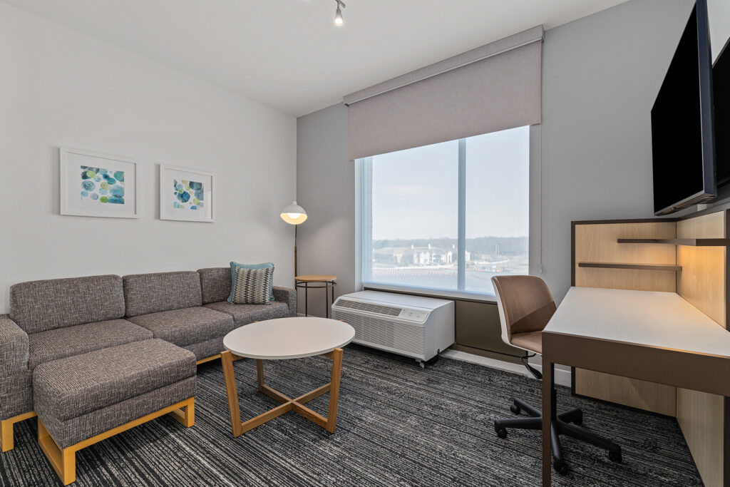 Modern TownePlace Suites hotel room in Murfreesboro, TN featuring a sectional sofa, round coffee table, desk and chair, large window with a shade, wall art, floor lamp, mounted TV, neutral tones, and a lovely view outside.