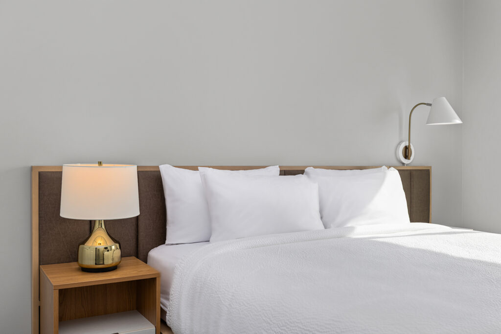 A neatly made bed with white linens and three pillows sits beside a wooden nightstand holding a gold lamp, under a wall-mounted white reading light, in a stylish Marriott hotel room in Murfreesboro against a plain light gray wall.